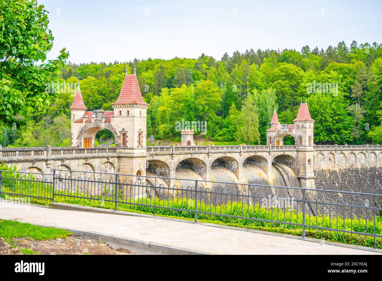 Ein Blick auf den Kralovstvi-Staudamm in Tschechien, mit dem berühmten burgähnlichen Gebäude mit seiner bogenförmigen Brücke. Der Damm ist von üppigem Grün umgeben und das Wasser fließt frei über den Überlauf. Stockfoto