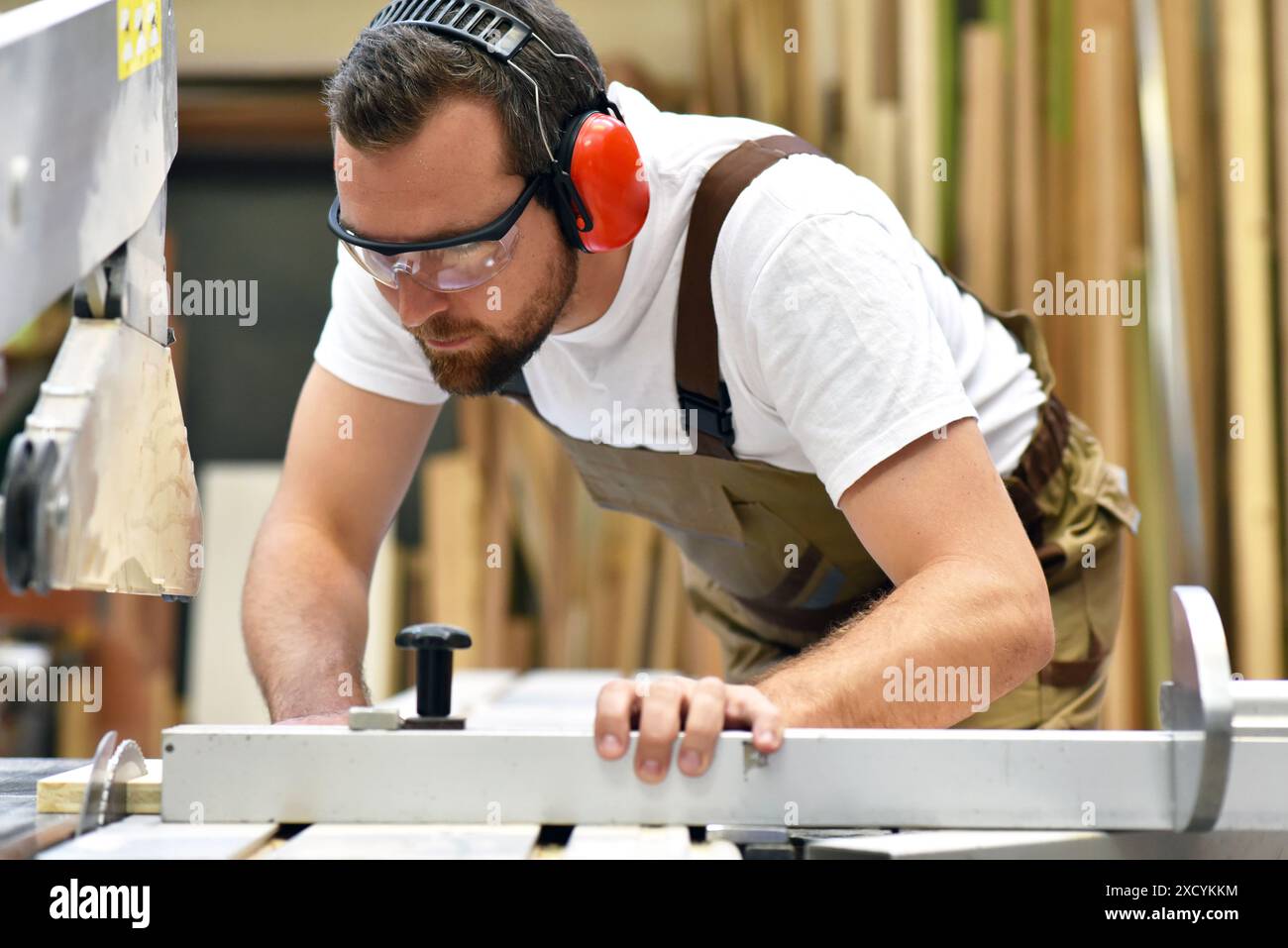 Junger Zimmermann in Arbeitskleidung arbeitet in der Schreinerei an einer Schleifmaschine - Arbeitskleidung mit Gehörschutz Stockfoto