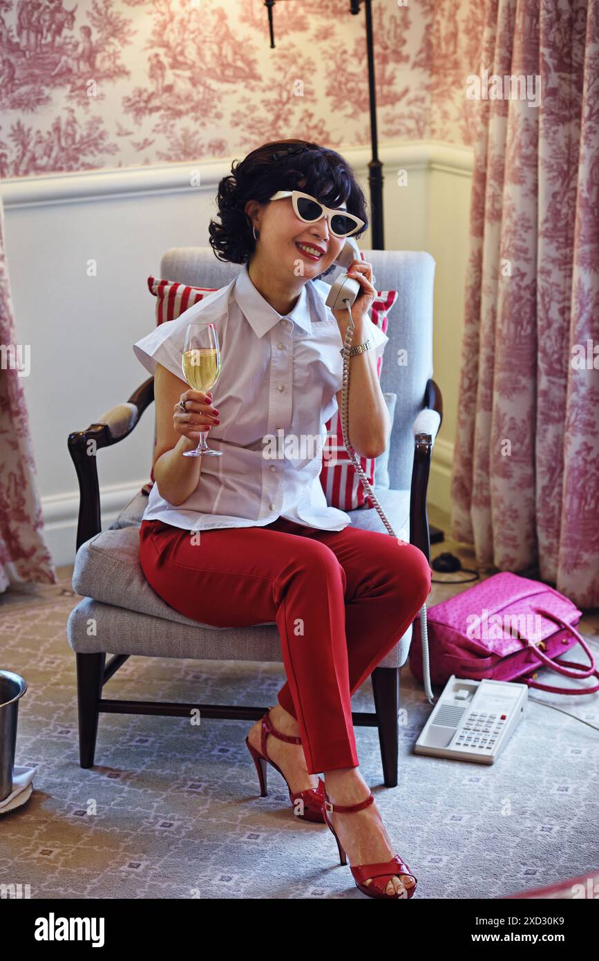 Retro-Frau aus den 1950er Jahren trinkt Champagner, während sie am Telefon teilnimmt, rote Skihosen, weiße Bluse und Sonnenbrille in einem hellen, gut gepolsterten Hotelzimmer Stockfoto