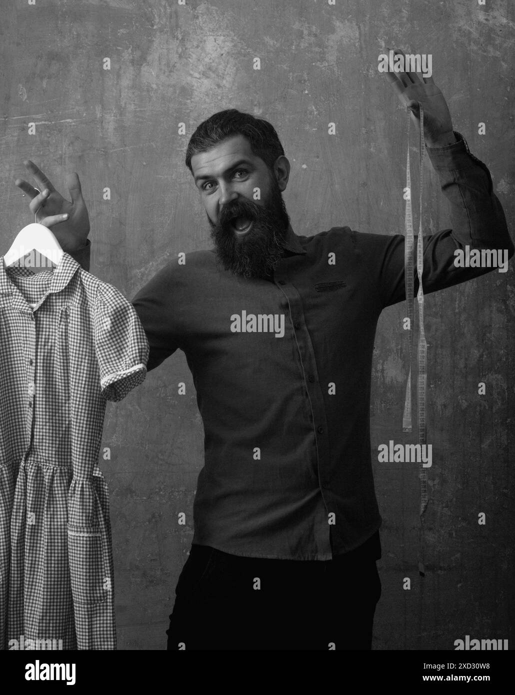 Lustiger aufgeregter bärtiger Mann näht Kleidung. Nähkleidung. Schneiderin hält Maß und weibliches Kleid. Mann Näherin. Die Schneiderin näht Kleidung für Frauen Stockfoto