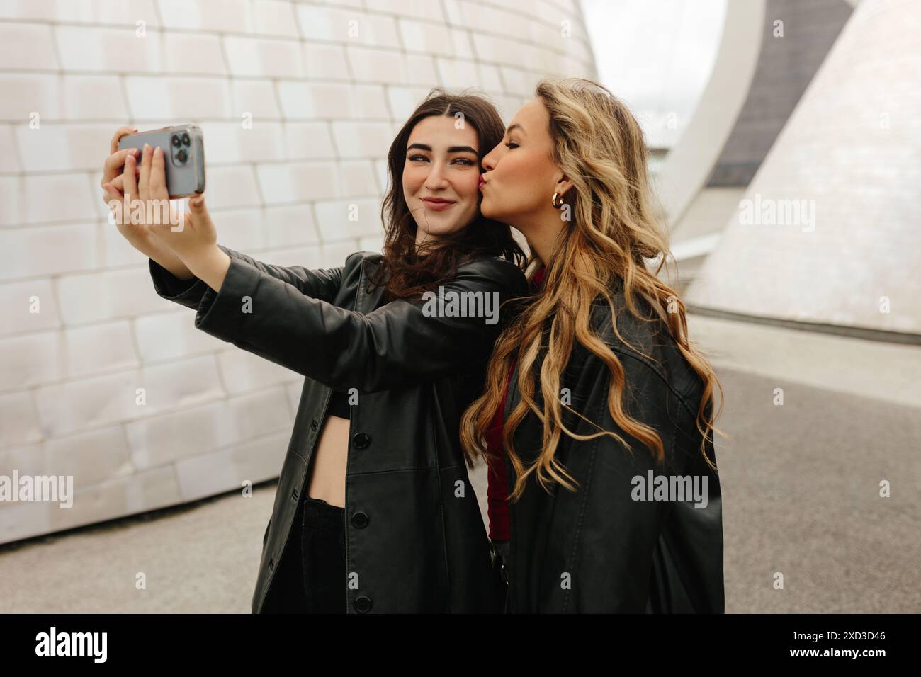 Zwei junge Frauen genießen einen Moment zusammen und machen ein Selfie mit einem Smartphone vor einem geometrischen, modernen Hintergrund. Einer küsst den anderen auf der che Stockfoto