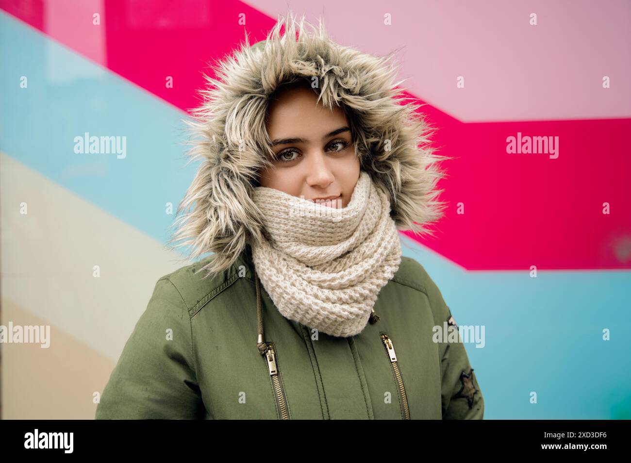 Eine junge Frau, die warme Winterkleidung trägt, darunter eine Kapuze mit Pelzbesatz und ein Stricktuch, lächelt sanft vor einem lebendigen, farbenfrohen geometrischen Hintergrund Stockfoto