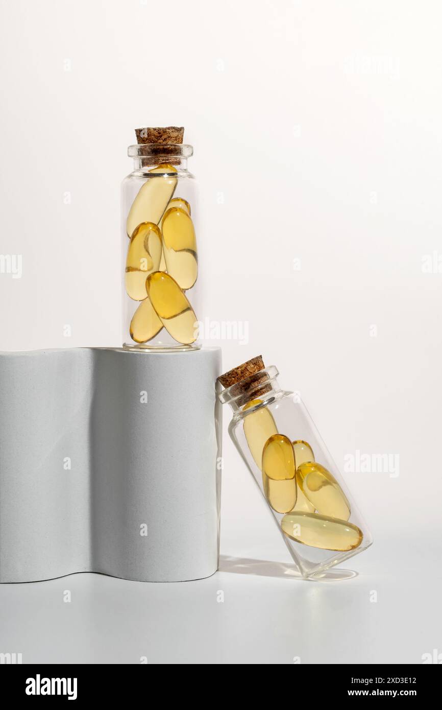 Zwei kleine Glasflaschen mit Korkdeckel, gefüllt mit gelben Omega-3-Kapseln, künstlerisch auf einem geschwungenen weißen Ständer gegen einen neutralen Hintergrund positioniert Stockfoto