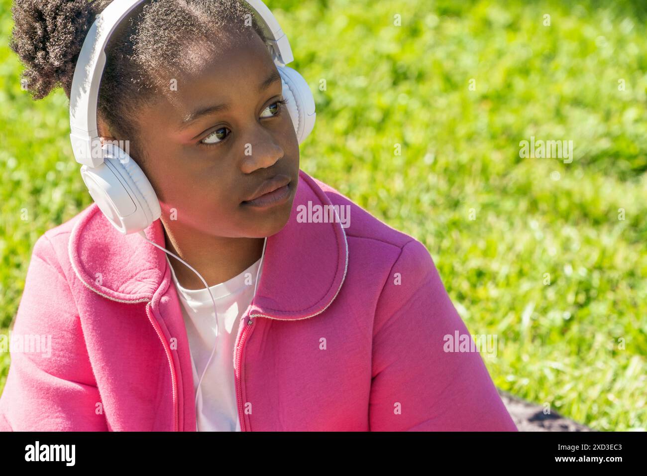 Ein junges schwarzes Mädchen sitzt in einem üppigen Park und genießt Musik über ihre weißen Kopfhörer Sie trägt eine leuchtend rosa Jacke über einem weißen Hemd Stockfoto