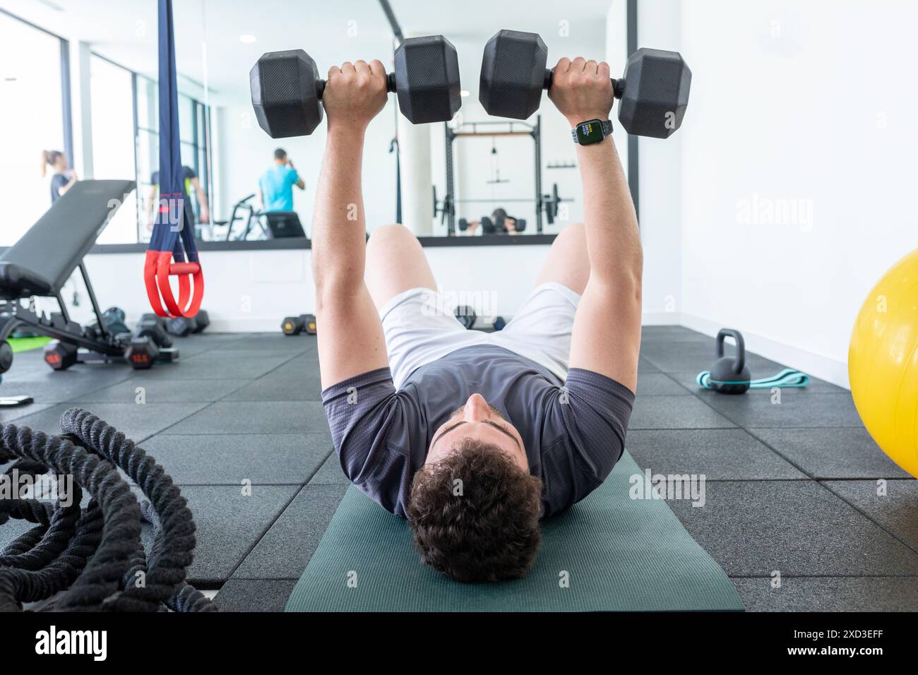 Ein junger Mann in sportlicher Kleidung liegt auf einer Matte in einem Fitnessstudio und konzentriert sich dabei, während er schwere schwarze Kurzhanteln für ein Brusttraining hebt, mit Trainingsgeräten und anderem Stockfoto