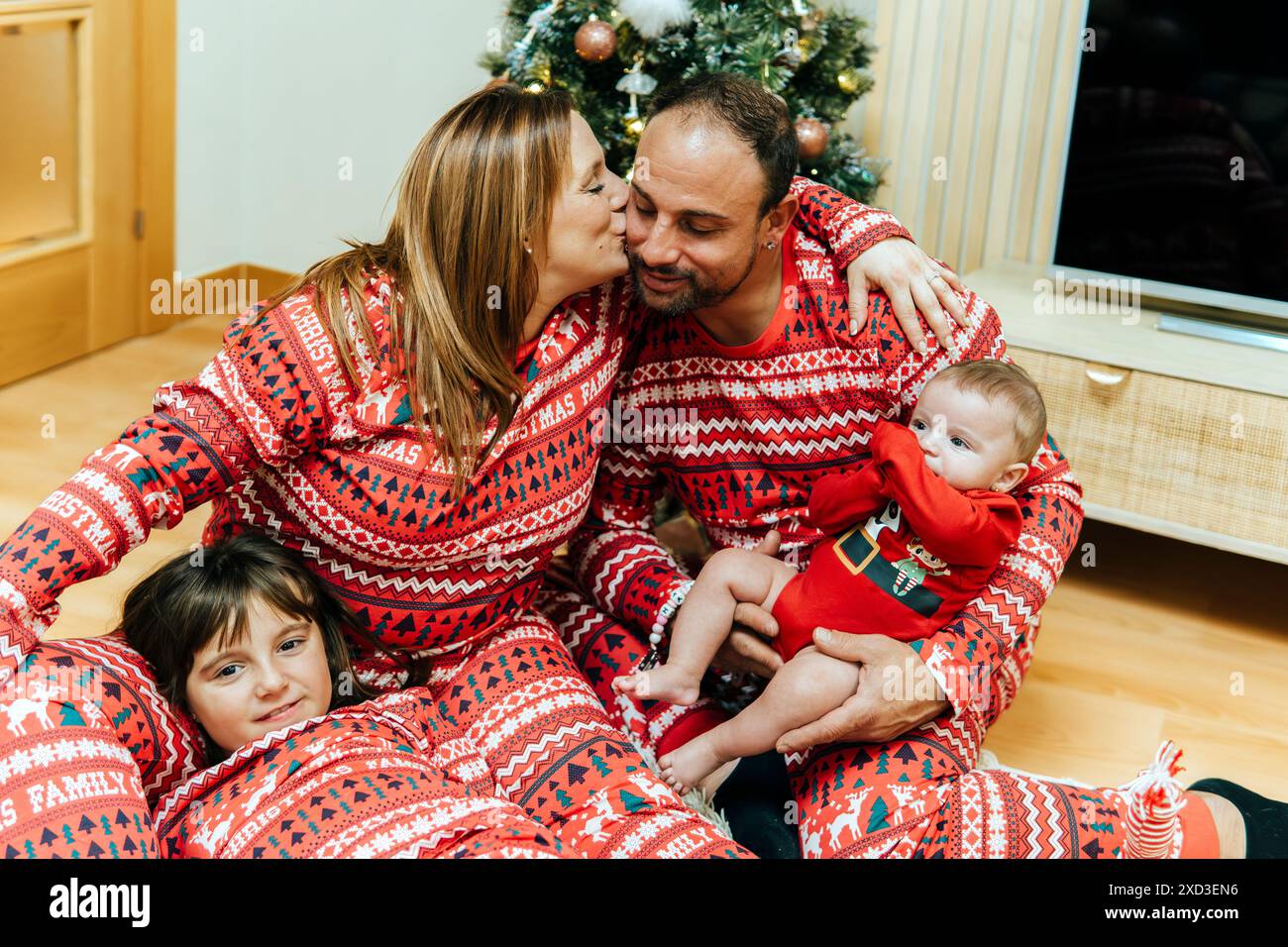 Vierköpfige Familie im passenden roten Weihnachts-Pyjama, die einen liebevollen Moment mit einem Neugeborenen teilt, neben einem verzierten Baum. Stockfoto