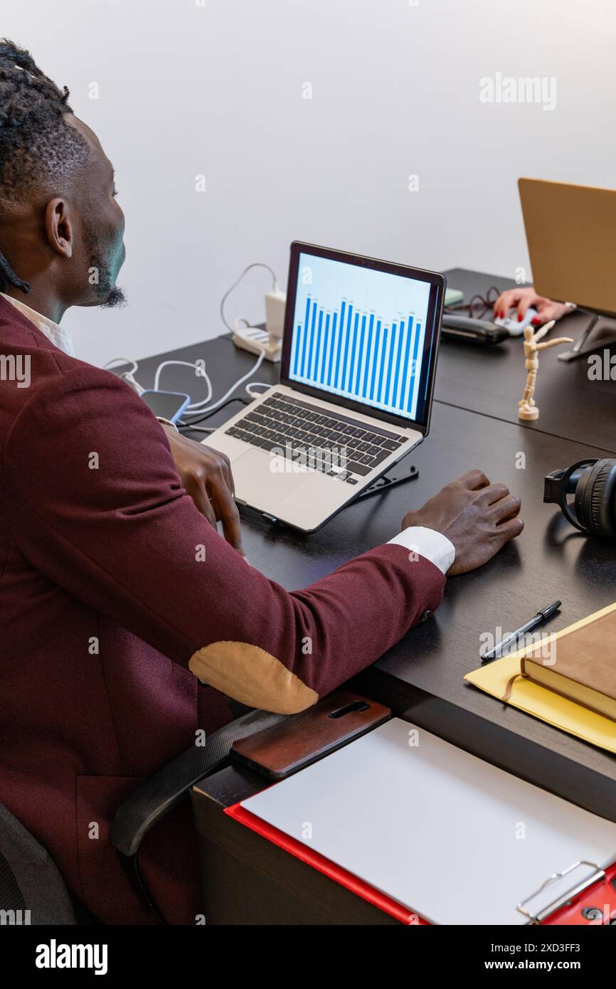 Ein fokussierter Geschäftsmann in einem braunen Anzug sieht Finanzdiagrammdaten auf einem Laptop an seinem Arbeitsplatz, umgeben von verschiedenen Büroartikeln und Gadgets Stockfoto