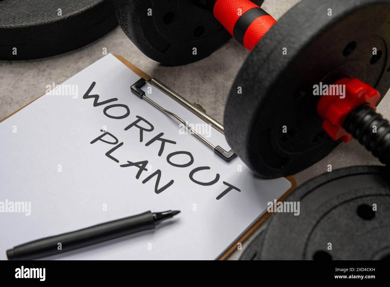 Ein Nahbild eines Clipboards mit einem TRAININGSPLAN, der neben einer schwarzen Hantel und Gewichtsplatten steht. Sport- und Fitnesskonzept. Stockfoto