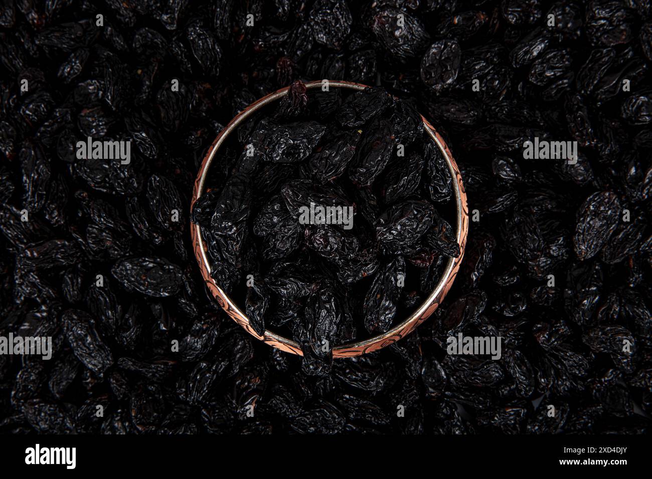 Schwarze Rosinen mit Samen (Sultaninen), getrocknete Früchte. Getrocknete schwarze Rosinen in einer Kupferschale, verstreute Rosinen um eine Kupferschale Rosinen. Stockfoto