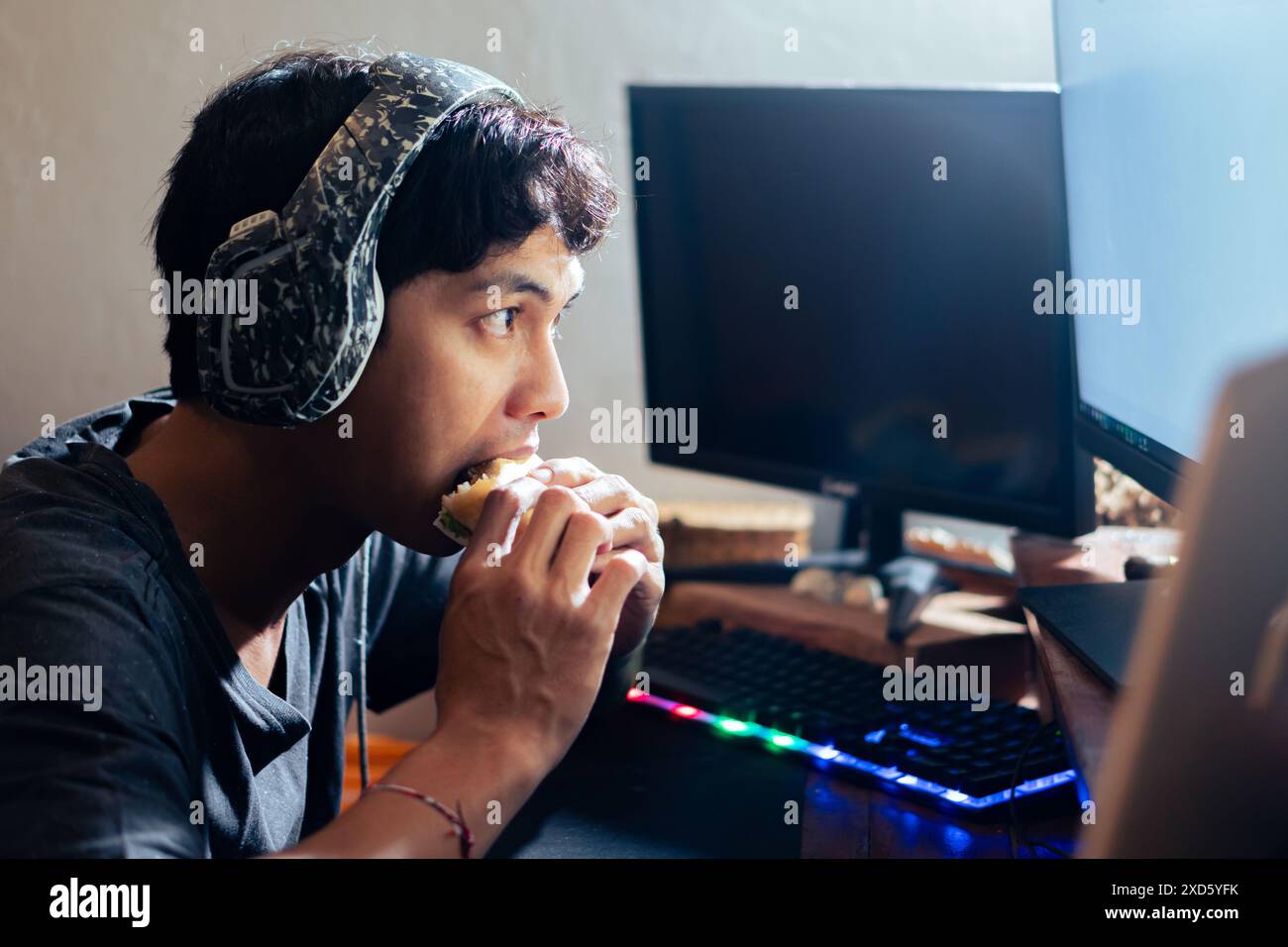 Junger Mann, der Kopfhörer trägt und ein Sandwich isst, während er am Computer arbeitet. Die Szene wird in einem schwach beleuchteten Raum mit zwei Monitoren gedreht. Stockfoto
