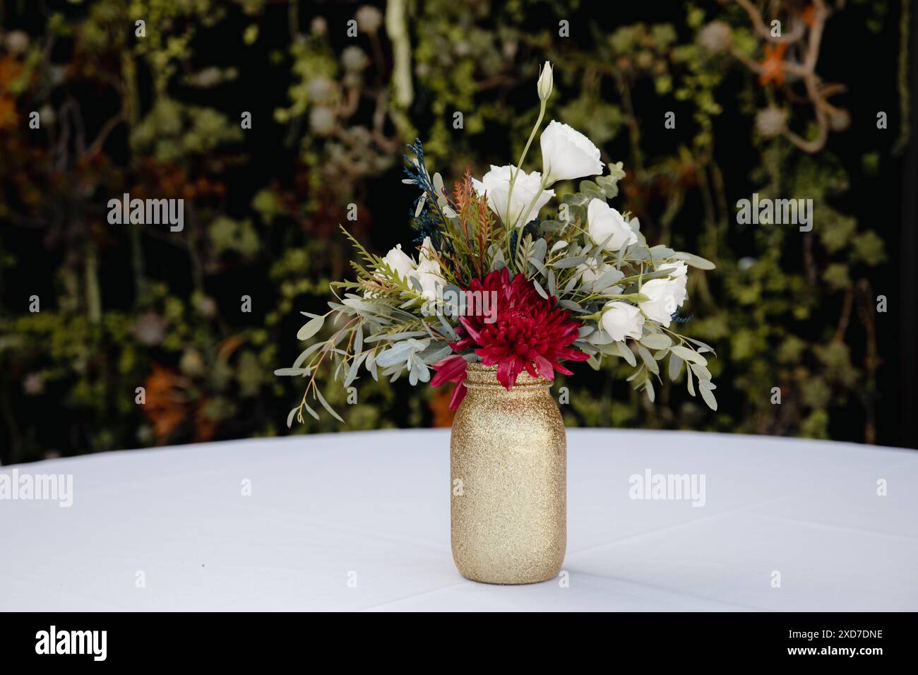 Hochzeitsempfang im Freien - Gäste-Tisch-Einrichtung. Goldene Glitzervase mit burgunderroten Pfingstrosen, Eukalyptusblätter Blumenarrangement Stockfoto
