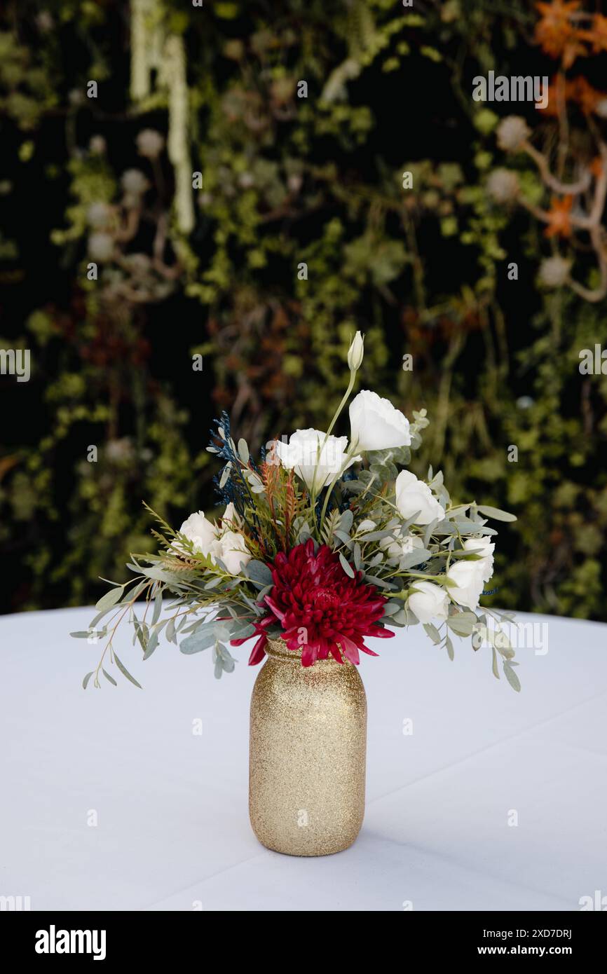 Hochzeitsempfang im Freien - Gäste-Tisch-Einrichtung. Goldene Glitzervase mit burgunderroten Pfingstrosen, Eukalyptusblätter Blumenarrangement Stockfoto