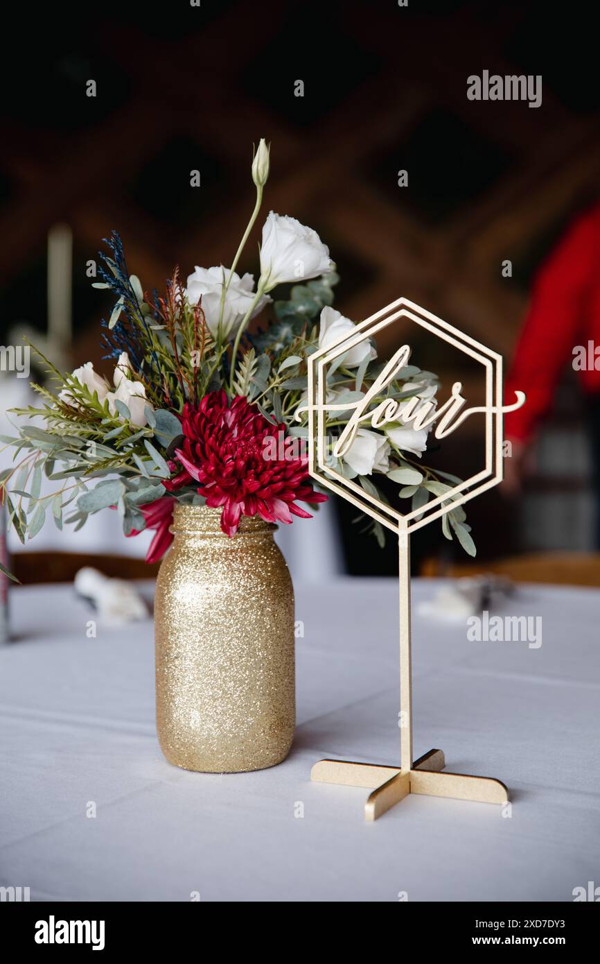 Hochzeitsempfang im Freien - Gäste-Tisch-Einrichtung. Goldene Glitzervase mit burgunderroten Pfingstrosen, Eukalyptusblätter Blumenarrangement Stockfoto
