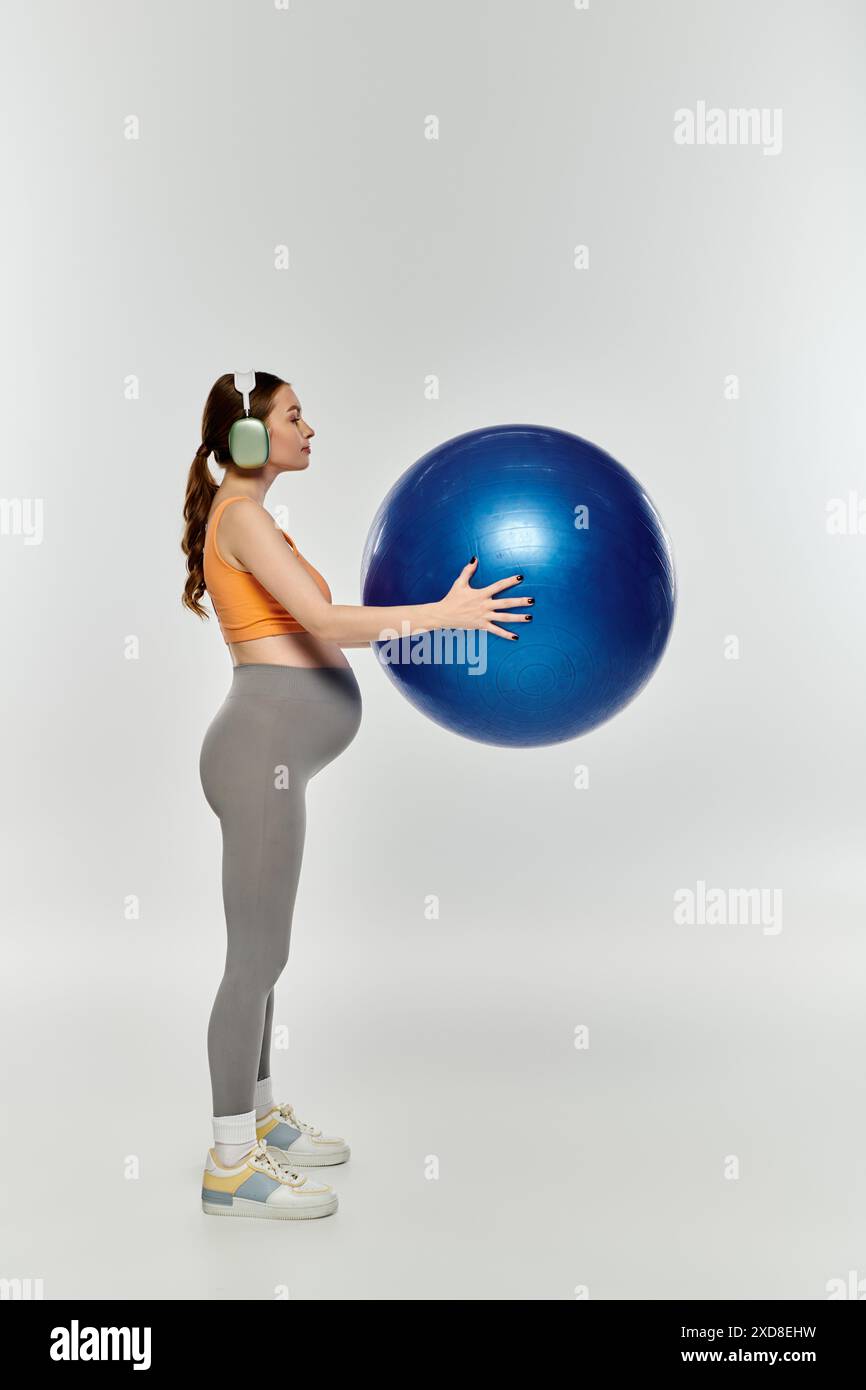 Eine junge, sportliche schwangere Frau in Aktivkleidung hält einen großen blauen Ball auf grauem Hintergrund. Stockfoto