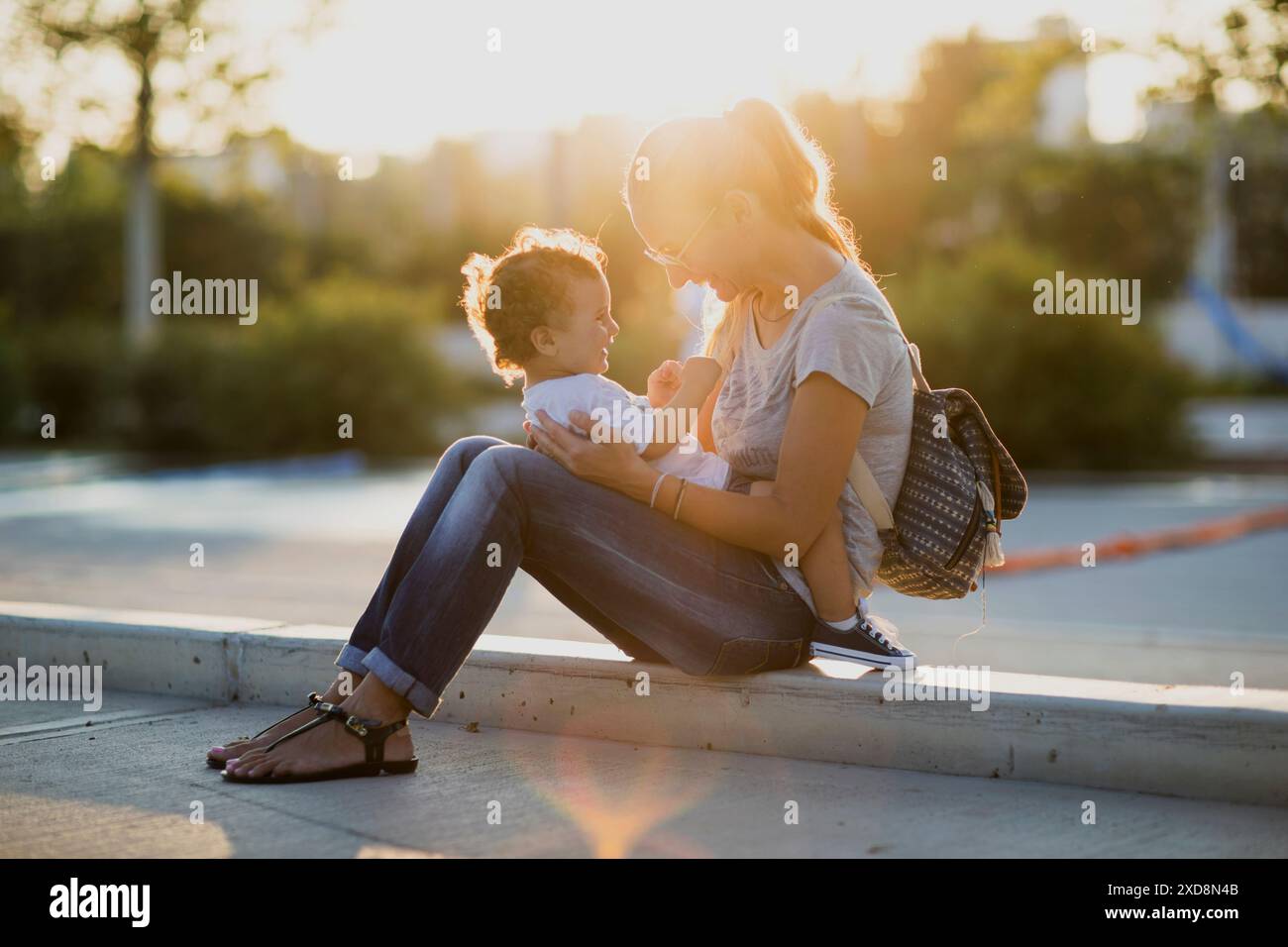 Eine Mutter sitzt mit ihrem kleinen Jungen auf einer Bordsteinkante und teilt einen Moment der Zärtlichkeit und Liebe, während das goldene Licht des Sonnenuntergangs sie taucht. Stockfoto