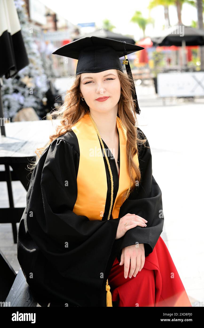 Weibliche Absolventin in Mütze und Kleid, sitzt draußen und sieht stolz aus Stockfoto
