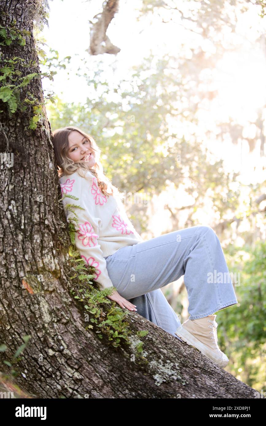 Trendige junge Frau mit welligen blonden Haaren, die auf einem Baumstamm sitzt Stockfoto
