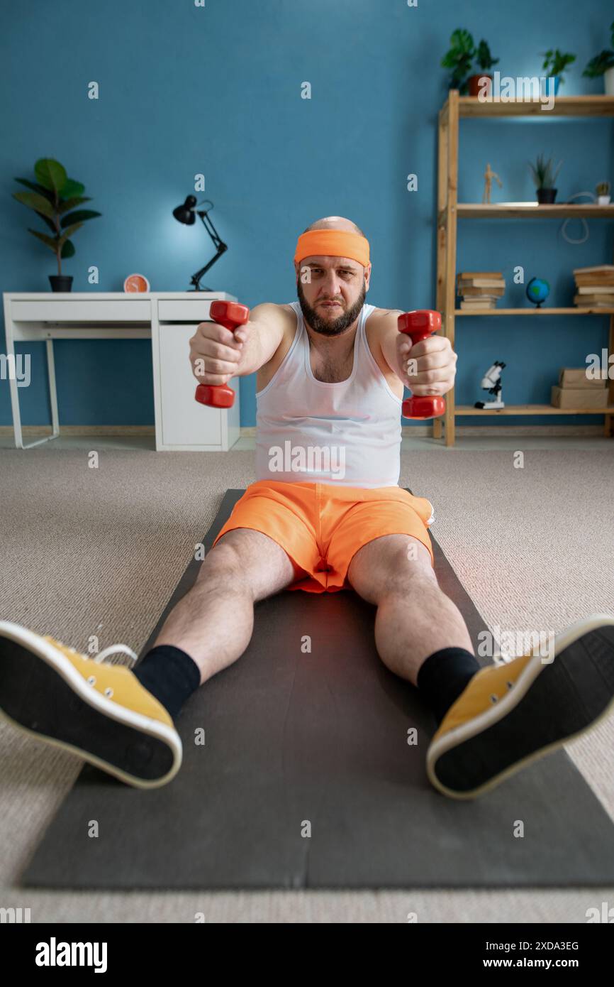 Lustiger Mann in orangen Shorts, der mit kleinen Kurzhanteln trainiert, die Bizepsparodie pumpen. Sport zu Hause Stockfoto