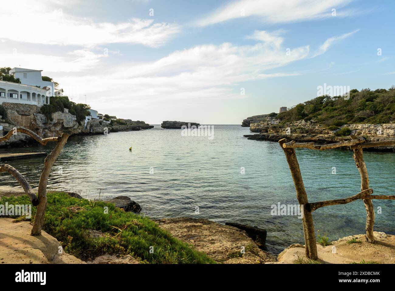 Ein ruhiger Blick auf die Bucht Cala Alfacar auf Menorca bei Sonnenuntergang mit ruhigem Wasser, weißen Gebäuden an den Klippen und üppigem Grün vor einem dramatischen Himmel. Stockfoto