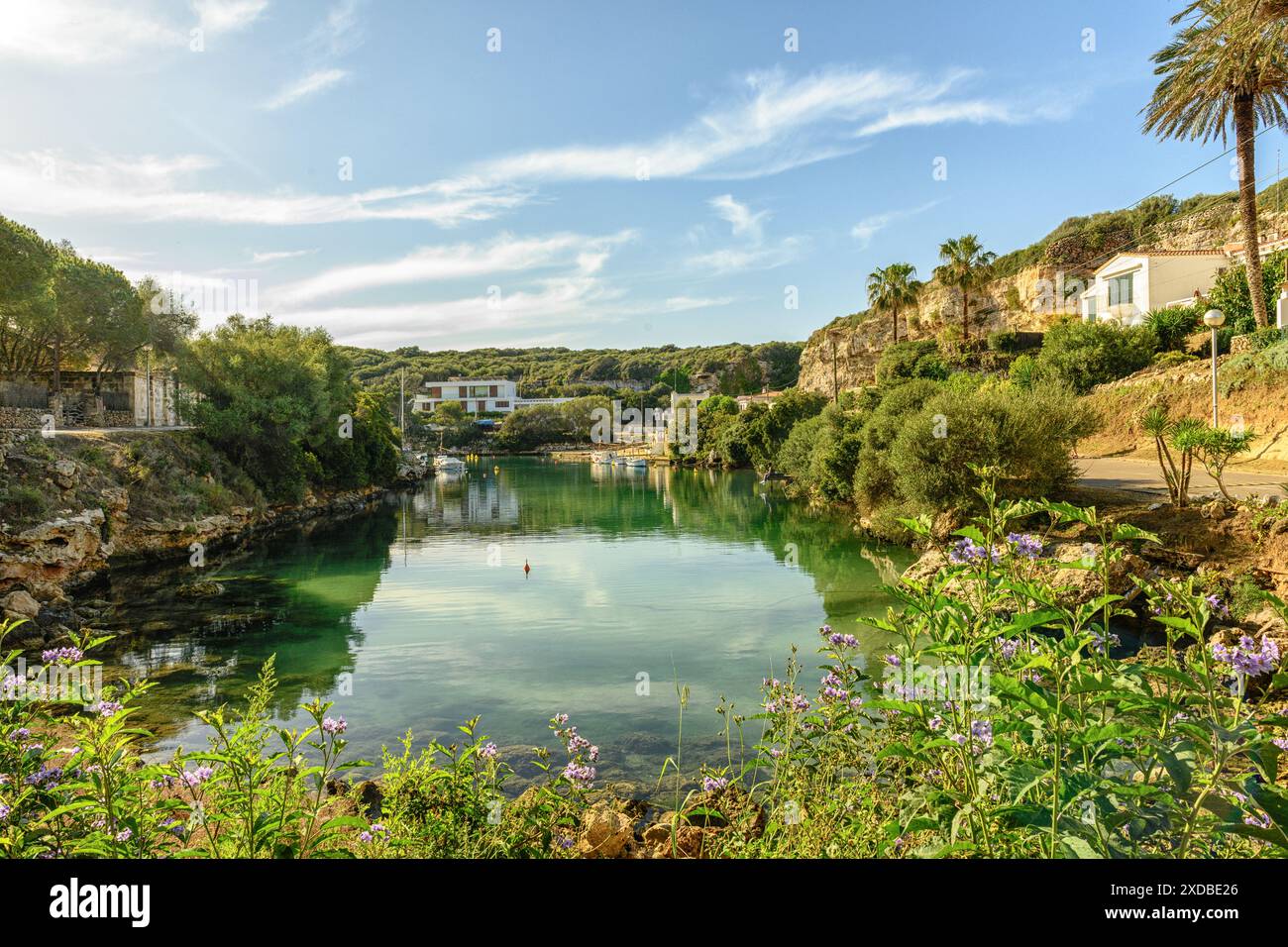 Ruhiger Hafen in Cala Sant Esteve auf Menorca, mit ankerbaren Booten, üppigem Grün und charmanten Häusern unter einem lebendigen, wolkenreichen Himmel. Stockfoto