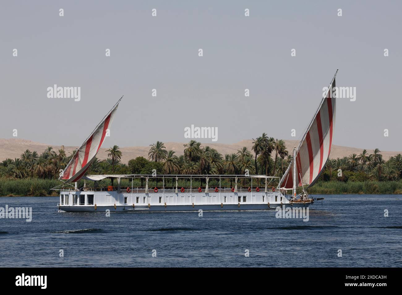 Ein Dahabiya-Segelboot mit zwei Masten und rot/weiß gestreiften Segeln fährt ruhig den Nil hinunter zwischen Luxor und Assuan in Ägypten. Stockfoto