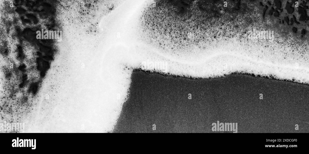 Ein Schwarzweiß-Foto, das die schwingenden schäumenden Wellen des Ozeans an einem Sandstrand zeigt. Das Bild erfasst ein dynamisches abstraktes Muster, das b erzeugt wurde Stockfoto