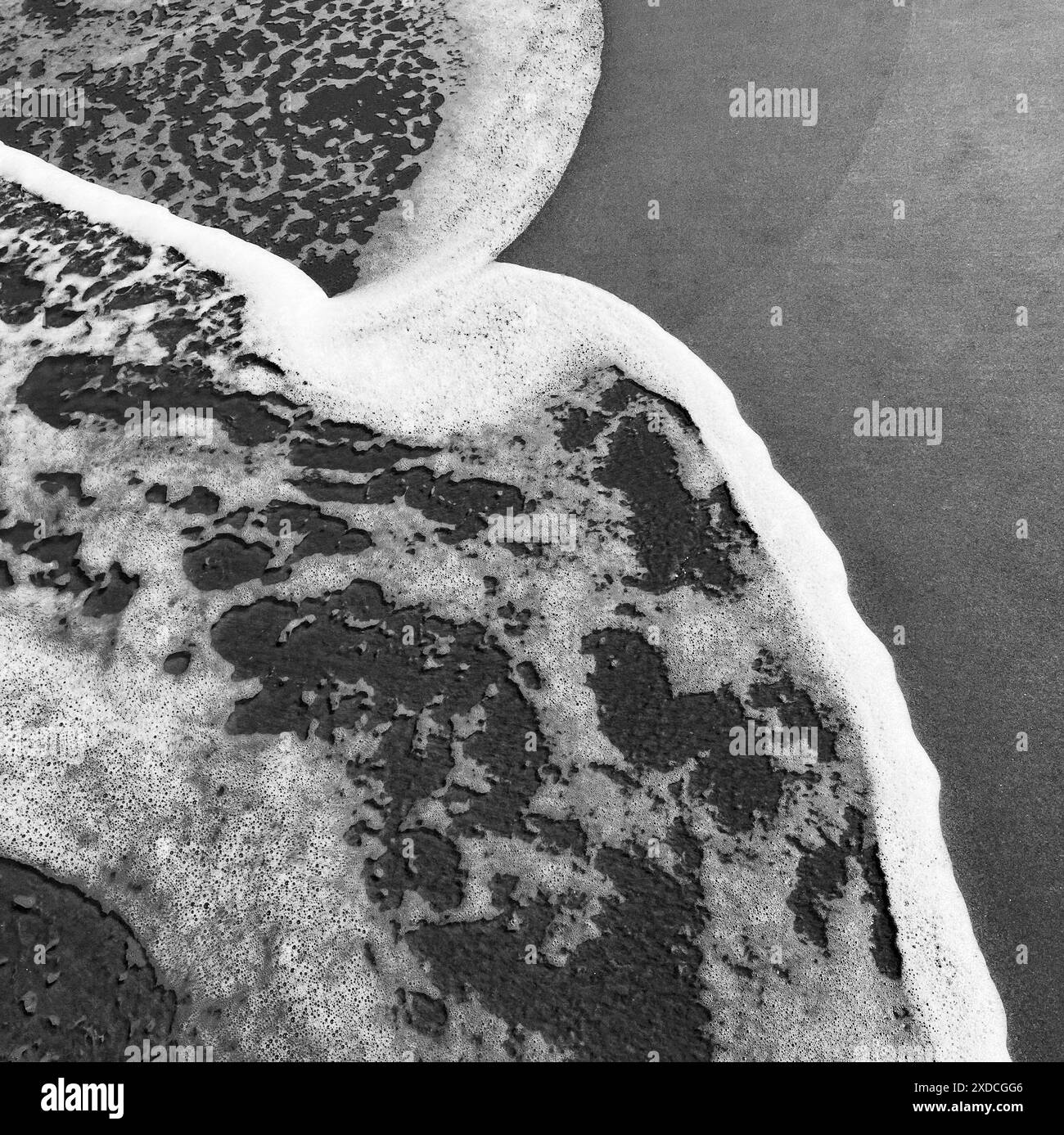 Ein Schwarzweiß-Foto, das die schwingenden schäumenden Wellen des Ozeans an einem Sandstrand zeigt. Das Bild erfasst ein dynamisches abstraktes Muster, das b erzeugt wurde Stockfoto