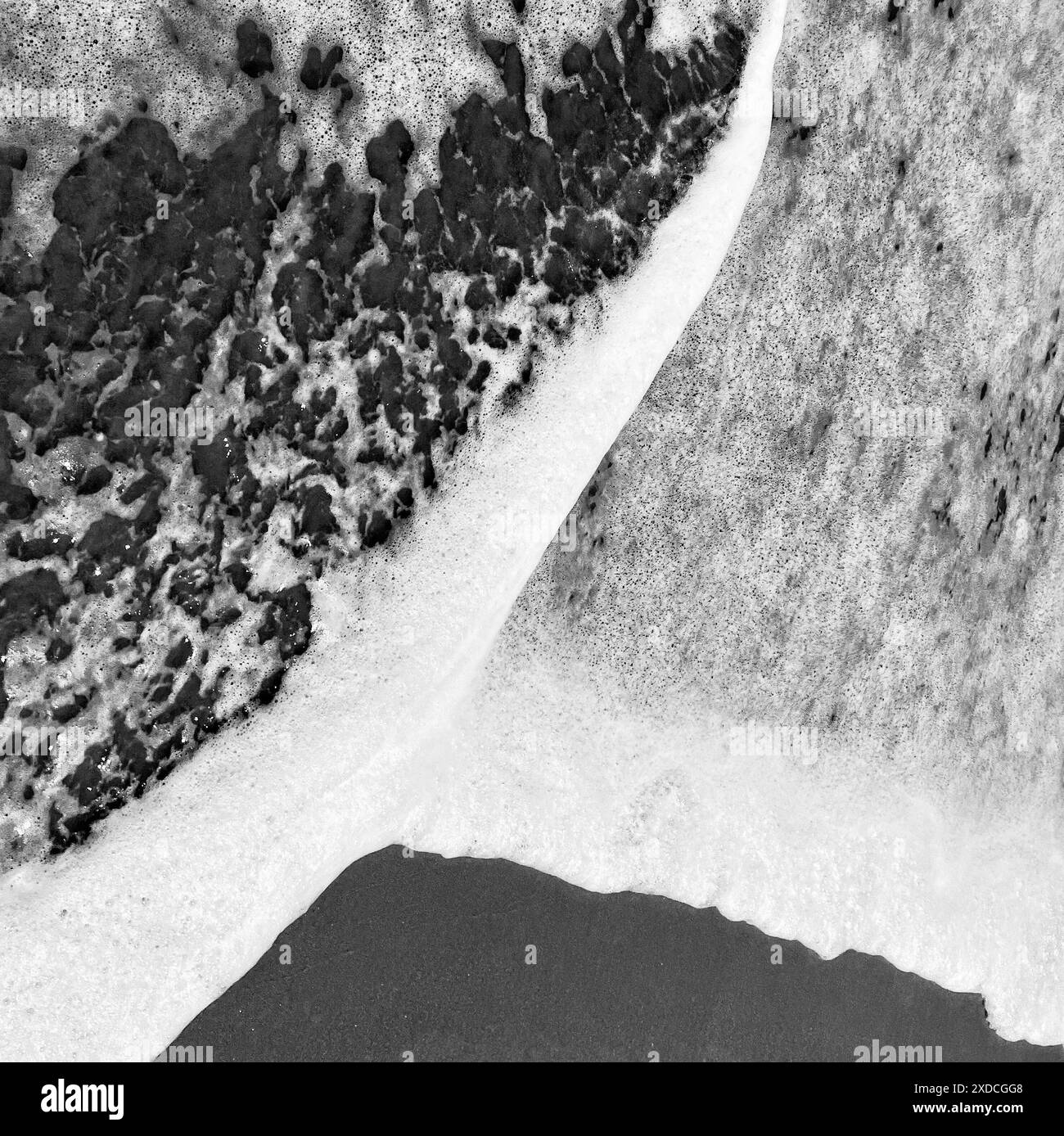 Ein Schwarzweiß-Foto, das die schwingenden schäumenden Wellen des Ozeans an einem Sandstrand zeigt. Das Bild erfasst ein dynamisches abstraktes Muster, das b erzeugt wurde Stockfoto