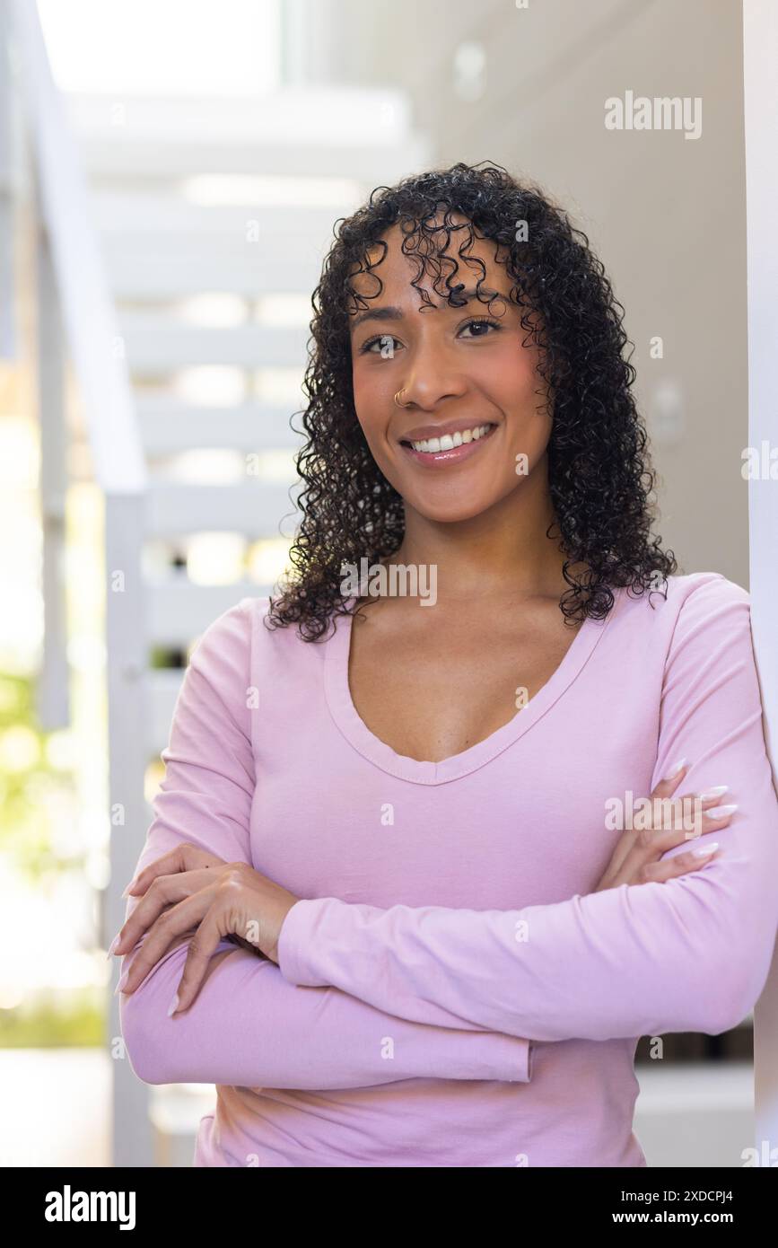 Lächelnde Frau mit lockigem Haar, die zu Hause mit gekreuzten Armen steht und selbstbewusst aussieht Stockfoto