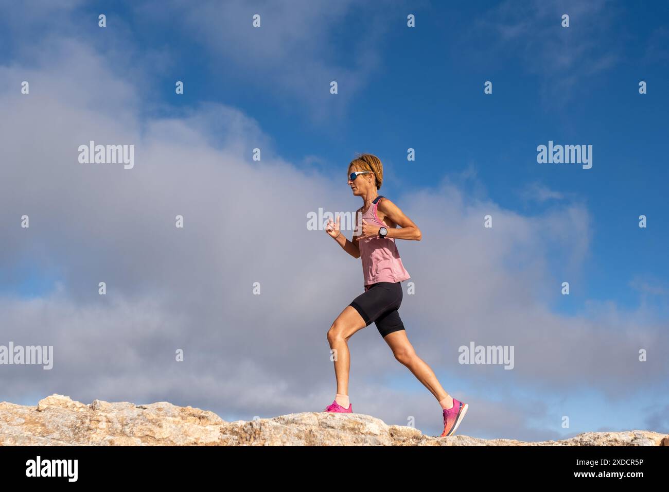 Eine athletische Frau läuft auf einem felsigen Hügel mit einem pinkfarbenen Hemd und schwarzen Shorts. Der Himmel ist blau mit ein paar Wolken. Sie ist entschlossen, den Gipfel zu erreichen Stockfoto