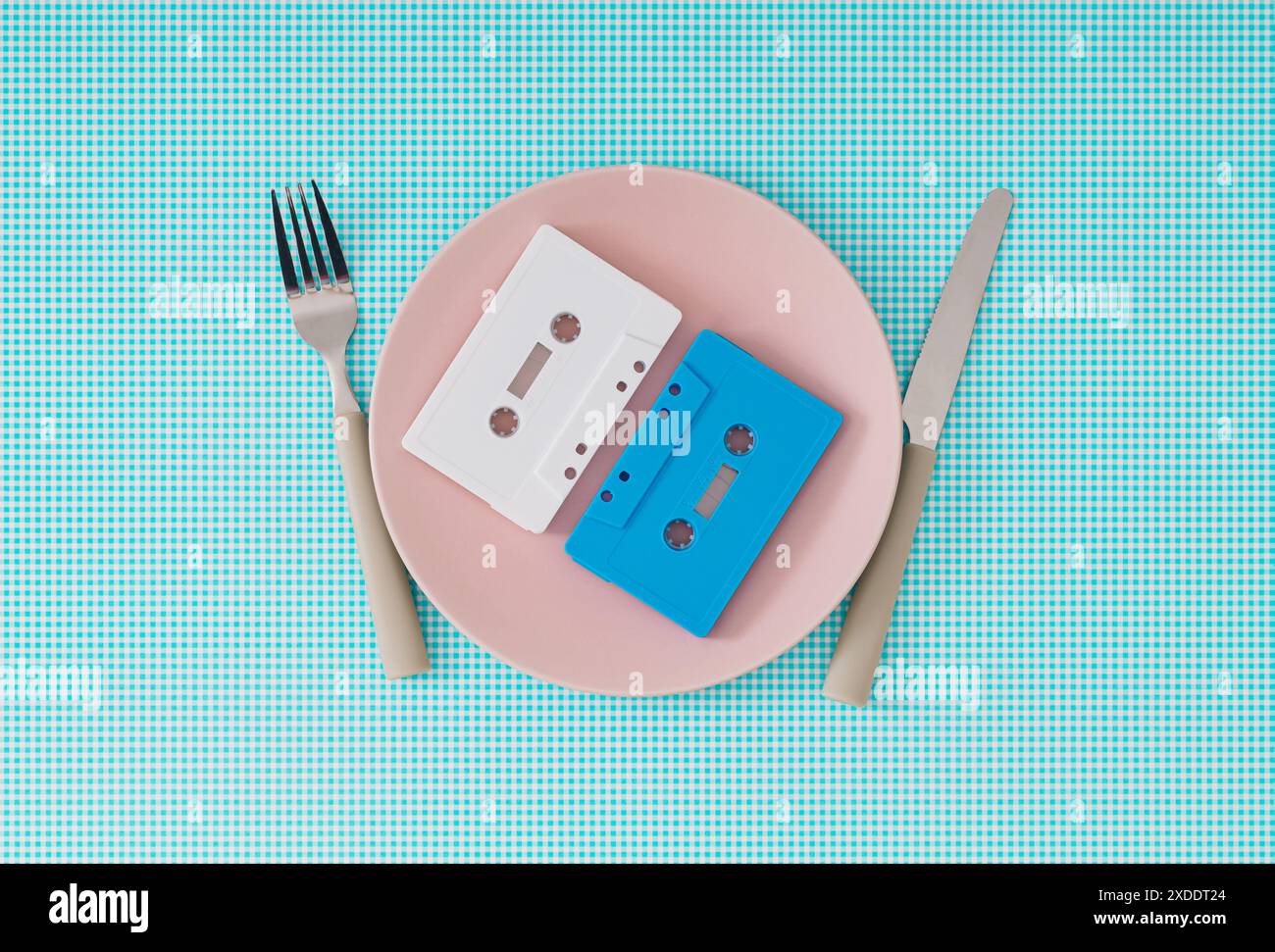 Audiokassettenbänder auf rosafarbener Platte mit Messer und Gabel auf weißem und blauem Hintergrund. Minimales Essen mit Retro-Musik-Idee. Stockfoto
