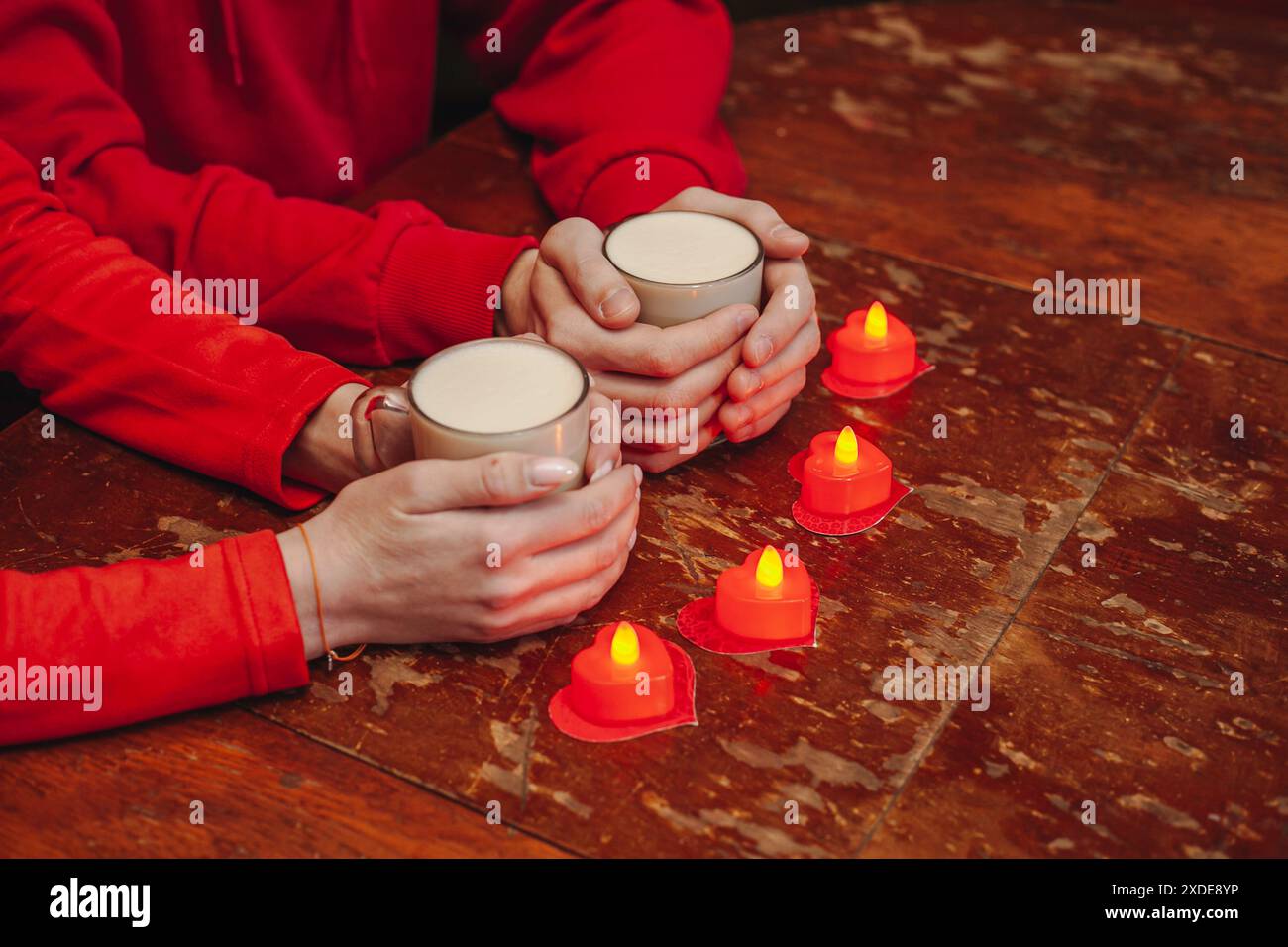 Eine Nahaufnahme eines Paares, das warme Getränke in der Hand hält, mit einer Reihe herzförmiger Kerzen auf einem Holztisch davor. Das Paar ist mit Stockfoto