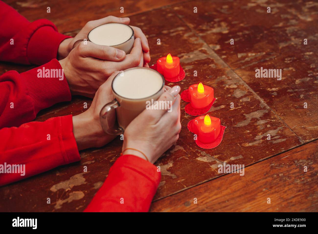 Eine Nahaufnahme eines Paares, das warme Getränke in der Hand hält, mit einer Reihe herzförmiger Kerzen auf einem Holztisch davor. Das Paar ist mit Stockfoto
