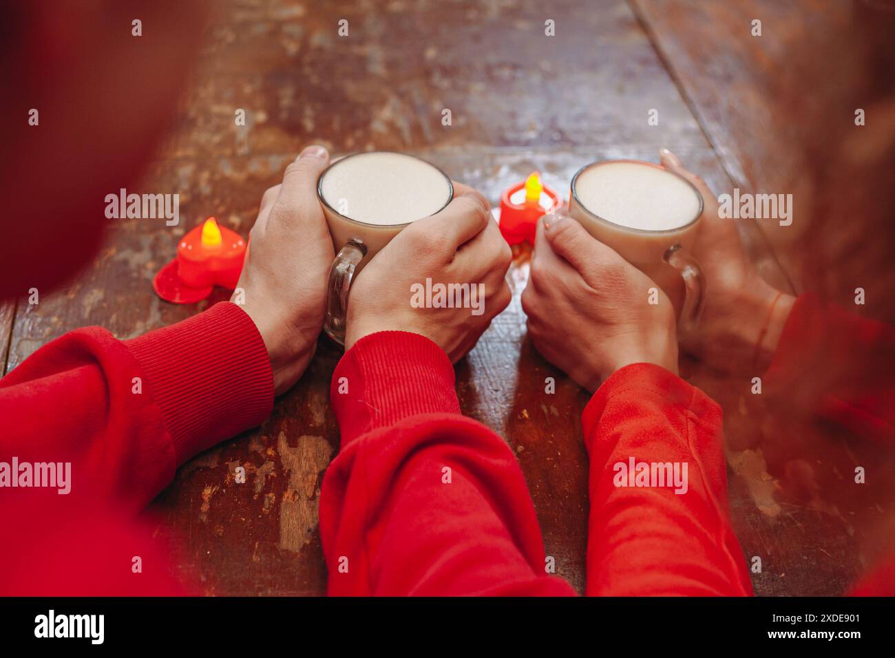 Eine Nahaufnahme eines Paares, das warme Getränke in der Hand hält, mit einer Reihe herzförmiger Kerzen auf einem Holztisch davor. Das Paar ist mit Stockfoto