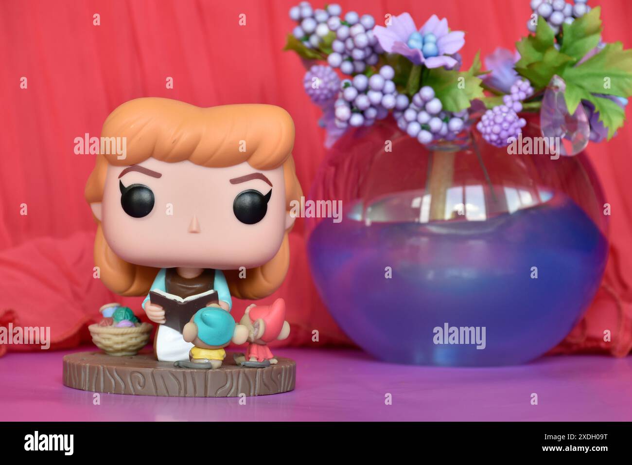 Funko Pop Actionfigur der Disney Prinzessin Cinderella im Brautjungfernkleid mit Buch. Rosa Vorhang, lila Blumen, Glaskugel, Dekor, bunt, Märchen. Stockfoto