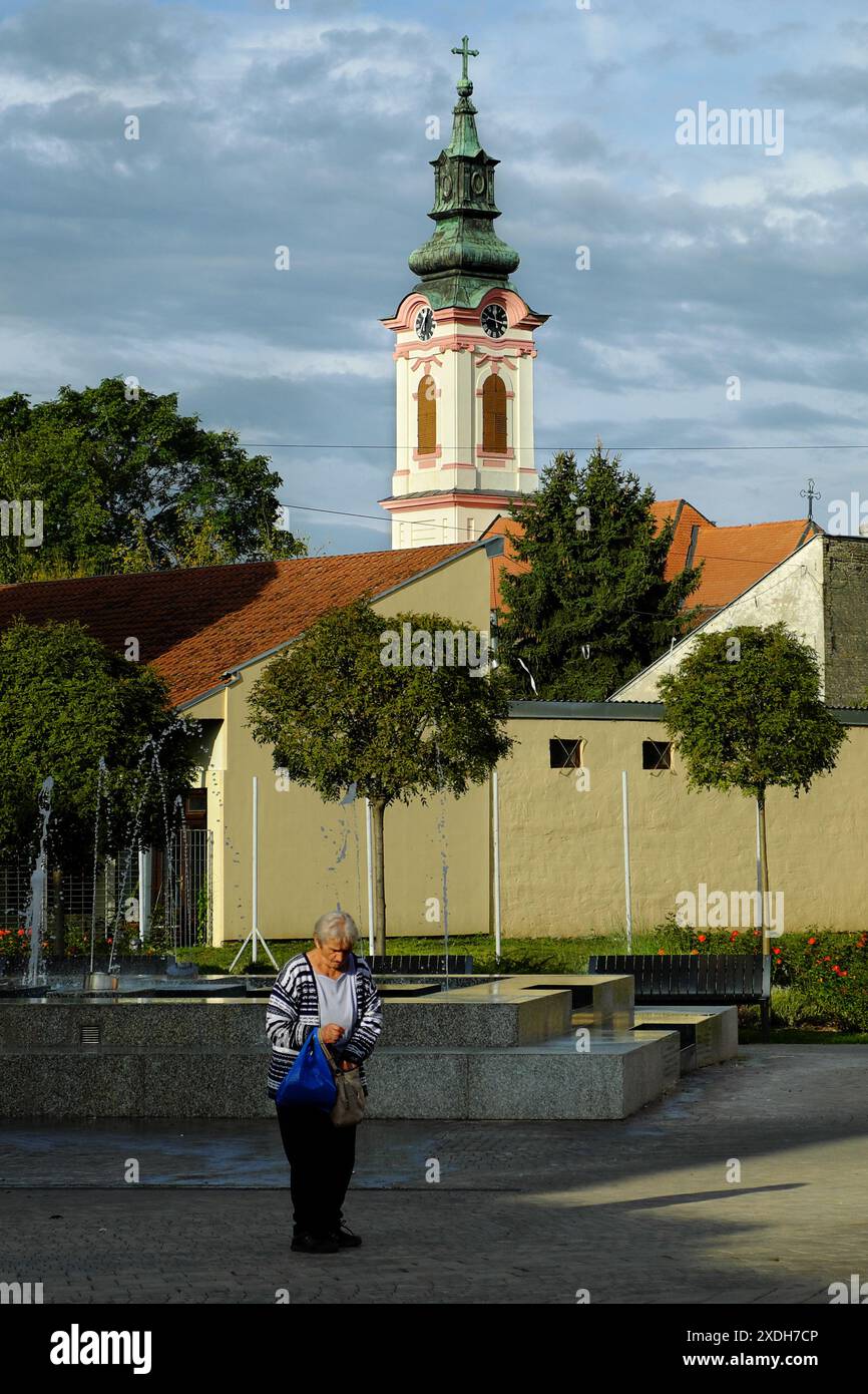 Alte Frau auf Hintergrund der Glockenturm der Kirche St. Georg in Sombor, Vojvodina, Serbien Stockfoto