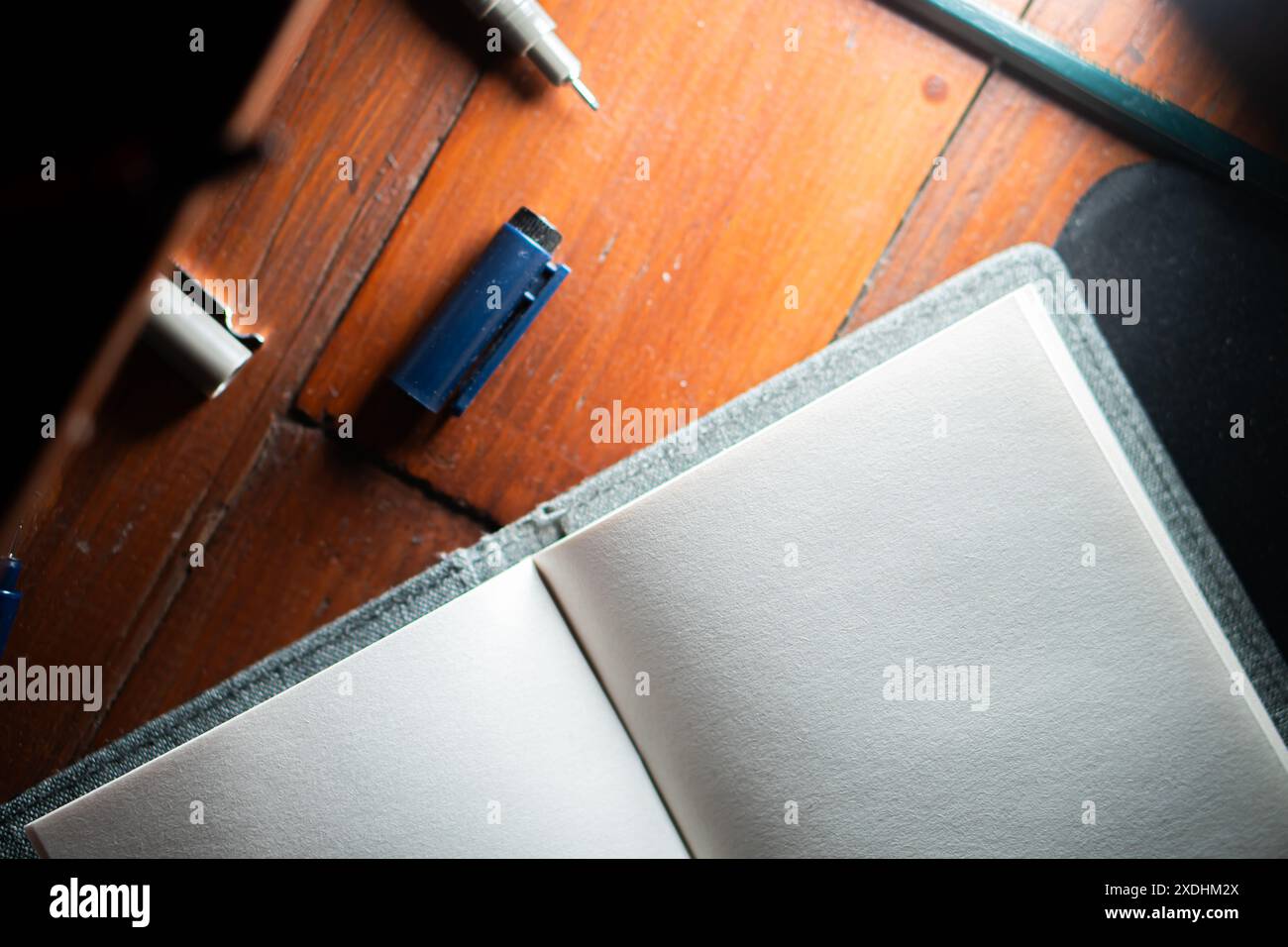 Ein offenes leeres Notizbuch auf einem Holzschreibtisch mit Stift, Bleistift und Spitzer in der Nähe. Die Szene ist gut beleuchtet und weist auf einen Arbeitsbereich hin, der zum Schreiben bereit ist Stockfoto