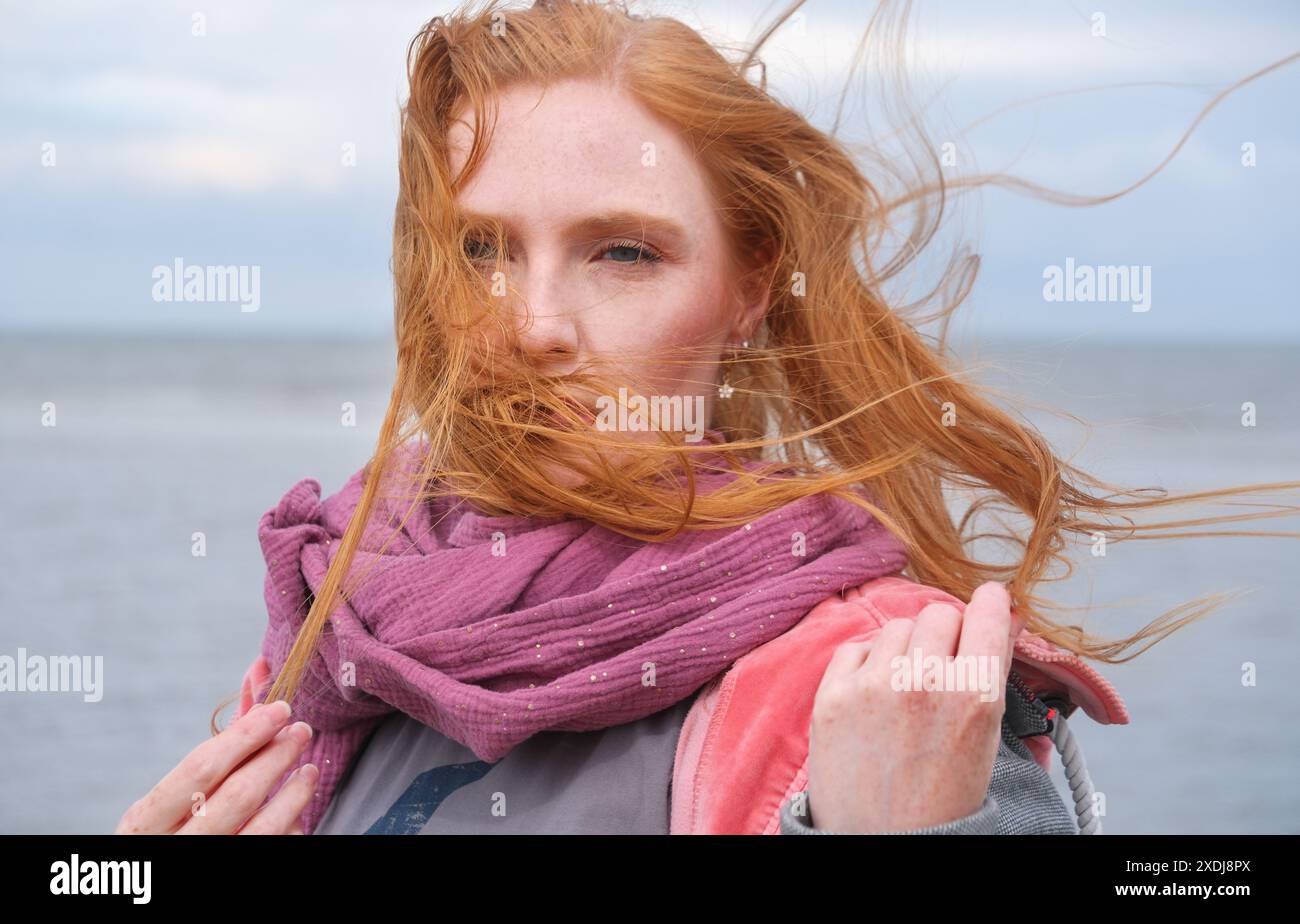 Eine Frau mit rotem Haar steht an einem windigen Tag am Meer, Haare wehen über ihr Gesicht, verdecken es teilweise, tragen einen lila Schal und eine rosa jacke Stockfoto