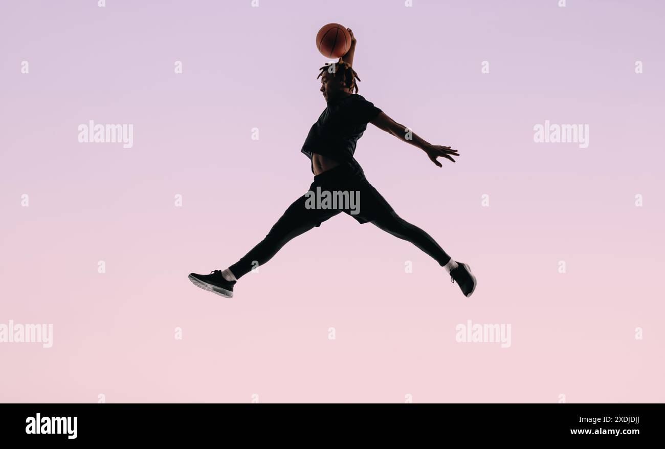 Passen Sie männliche Basketballspieler mit Dreadlocks, die während eines Spiels den Ball abtauchen. Seine Silhouette hebt sich vor einem rosafarbenen Hintergrund ab und zeigt sein A Stockfoto