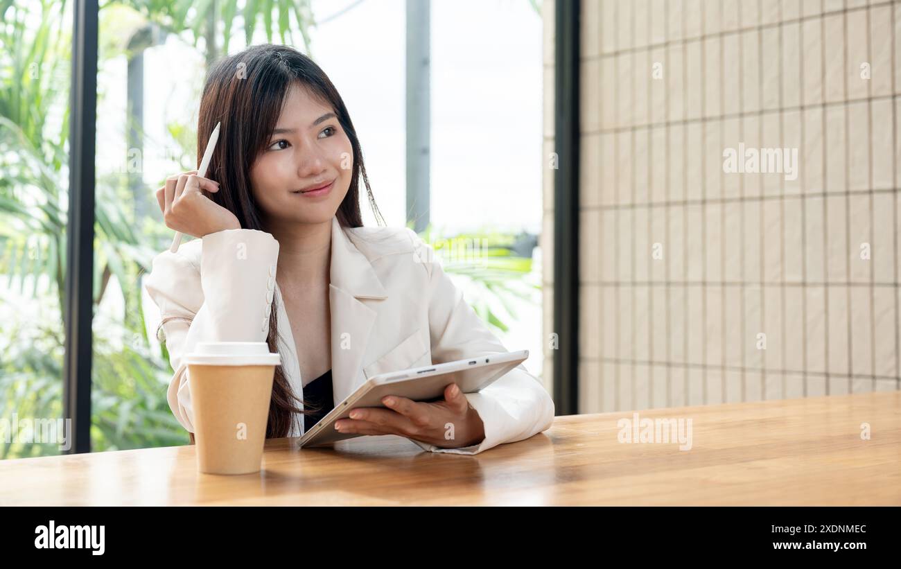 Eine attraktive, nachdenkliche asiatische Geschäftsfrau arbeitet von einem Café aus, verwendet ihr digitales Tablet, blickt auf die Außenansicht und denkt abou Stockfoto