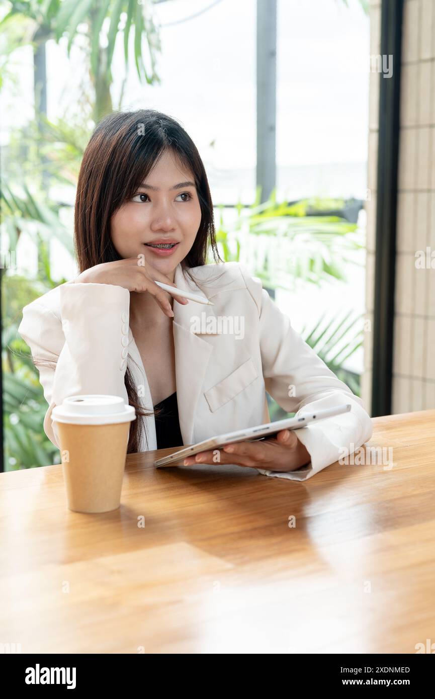 Eine attraktive, nachdenkliche asiatische Geschäftsfrau arbeitet von einem Café aus, verwendet ihr digitales Tablet, blickt auf die Außenansicht und denkt abou Stockfoto