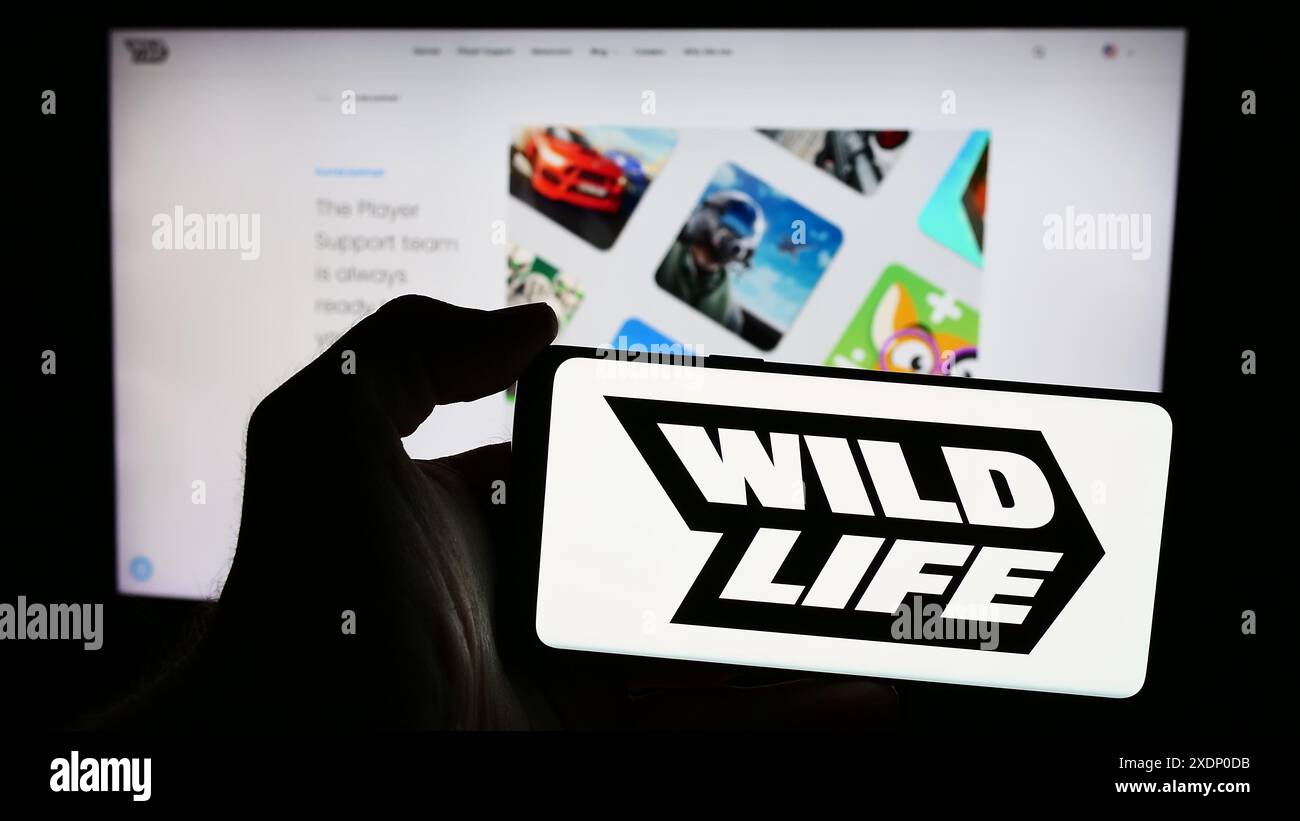 Person mit Mobiltelefon und Logo des brasilianischen Spieleunternehmens Wild Life Inc. (Wildlife Studios) vor der Webseite. Schwerpunkt auf dem Display des Telefons. Stockfoto