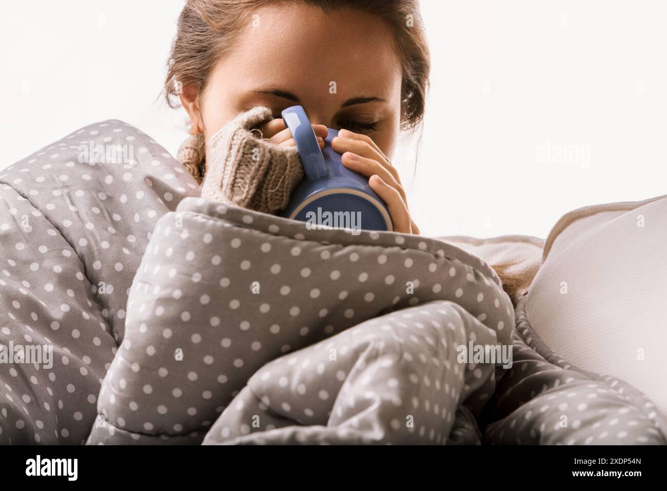 Müde Frau kuschelt sich unter eine Decke und schlürft ein heißes Getränk für Komfort zu Hause. Im Winter sucht sie Ruhe und Erholung in ihrem Wohnzimmer Stockfoto