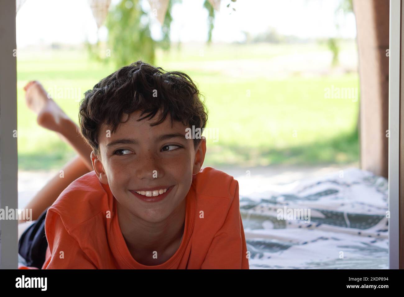 Junge, der in einem Wohnmobil-Bett mit orangefarbenem T-Shirt und Vegetation draußen liegt Stockfoto