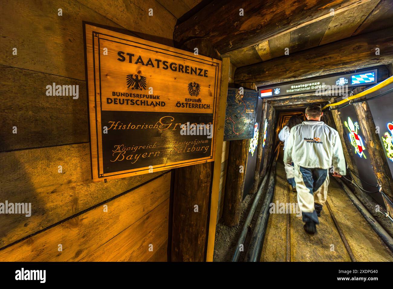 Ein Besucher in einer weißen Salzwelten-Jacke geht zurück zum Ausgang durch den Grenztunnel von Salzwelten Hallein bei Hallein, Salzburg. Ein Schild an der Wand erklärt die „Staatsgrenze“ zwischen Deutschland und Österreich sowie die historische Grenze zwischen dem Königreich Bayern und dem Erzbistum Salzburg. Die Grubenführung überquert die unterirdische Staatsgrenze einmal auf dem Hinweg und einmal auf dem Rückweg. Stockfoto