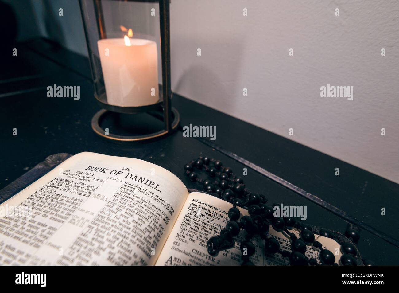 Eine Bibel, offen im Buch Daniel, auf einem hölzernen Sideboard mit schwarzen Rosenkranzperlen und einer Kerze dahinter Stockfoto