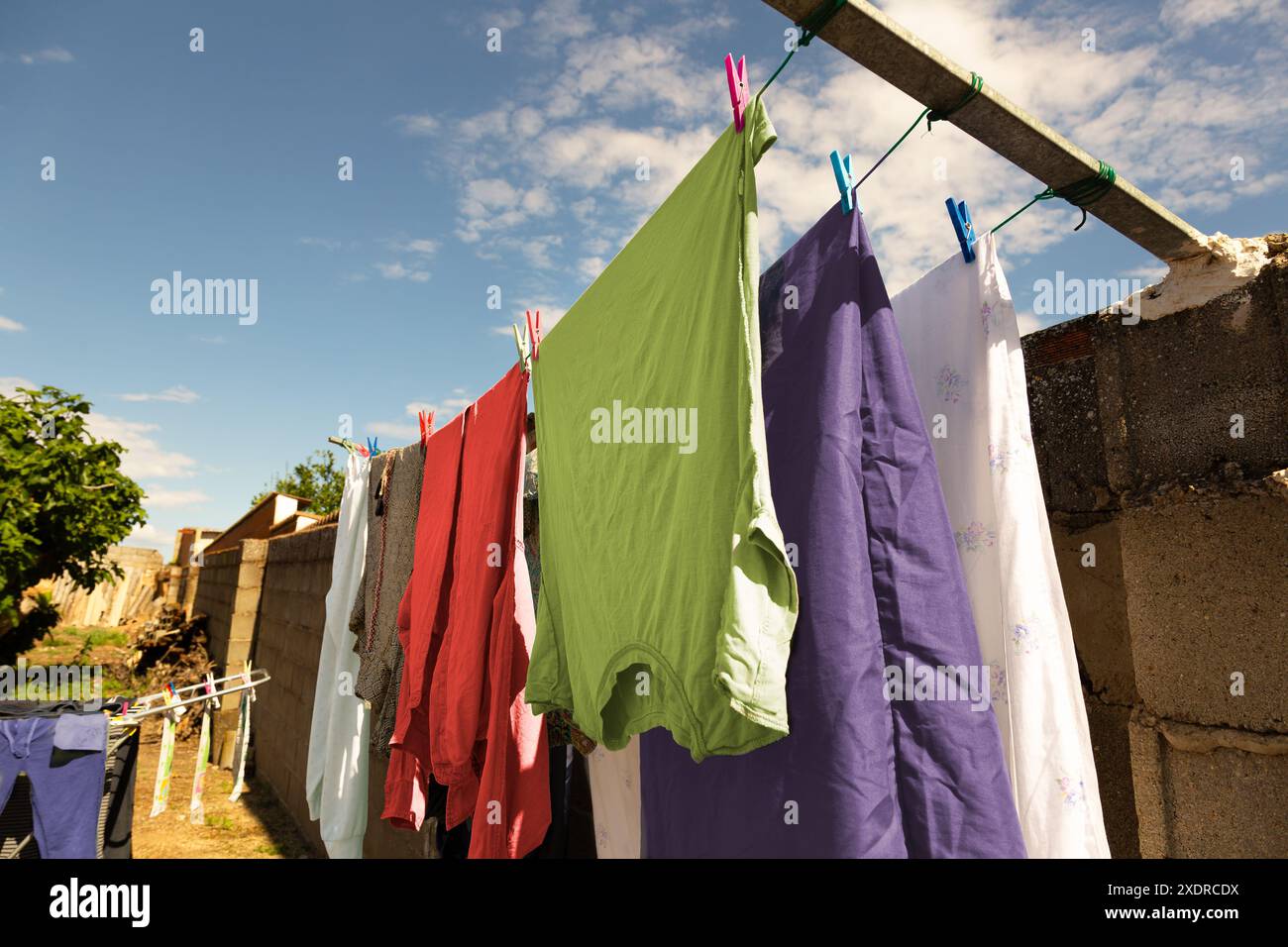 Farbenfrohe Kleidung hängt an einigen Seilen mit Kleiderschalen, im Innenhof des Dorfhauses, um an einem sonnigen Tag an der Luft zu trocknen, mit einigen Wolken im Blau Stockfoto