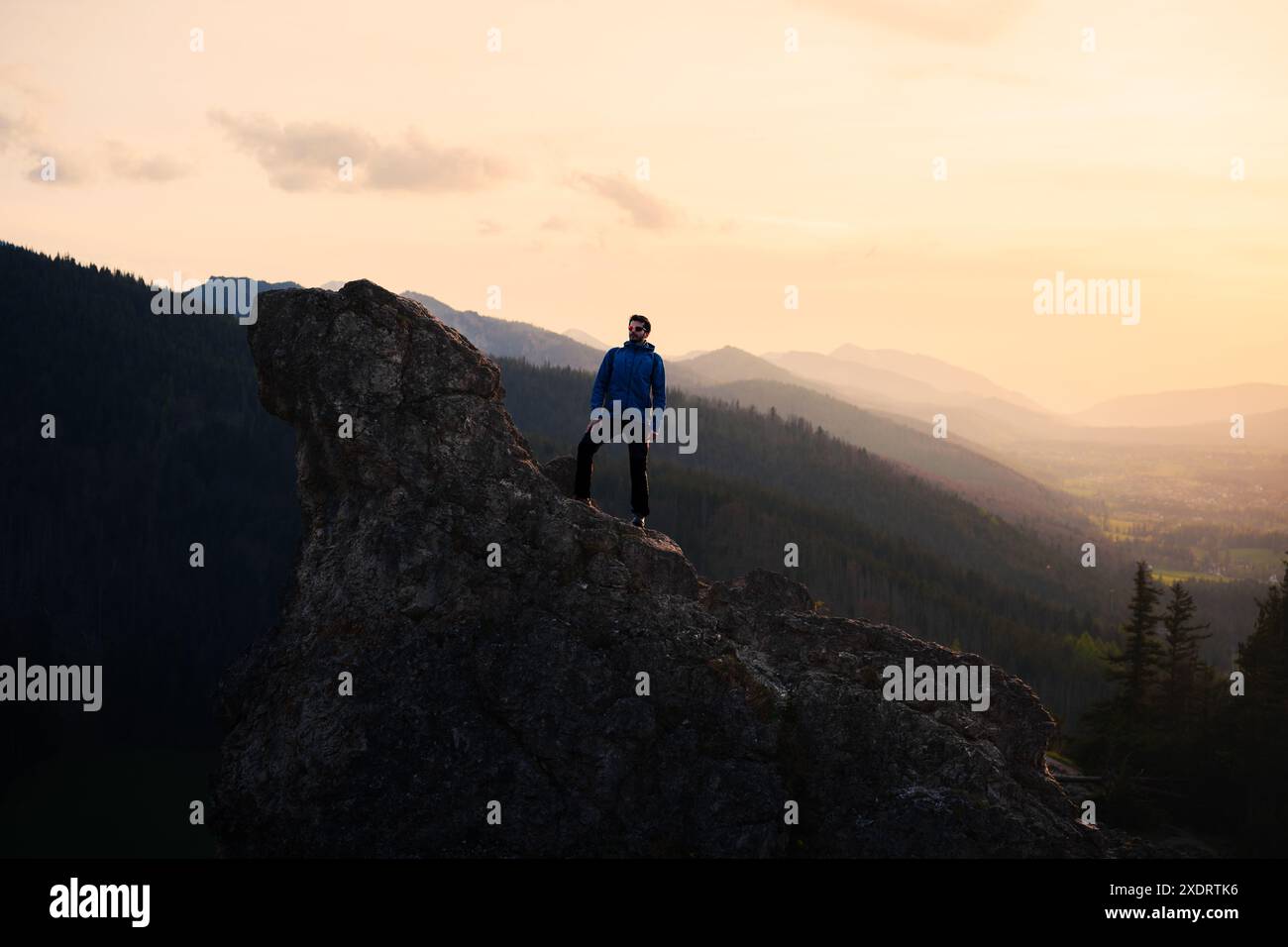 Ein Mann sitzt auf einem Felsen im Hochland, umgeben von Bergketten, während die Sonne untergeht, und erzeugt ein atemberaubendes atmosphärisches Phänomen am Himmel Stockfoto