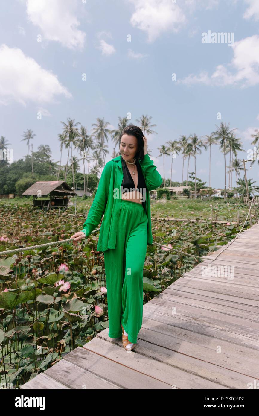 Schöne Frau in grünem Outfit spaziert entlang einer Holzbrücke über einen Seerosenteich, umgeben von Palmen und üppiger Vegetation. Stockfoto