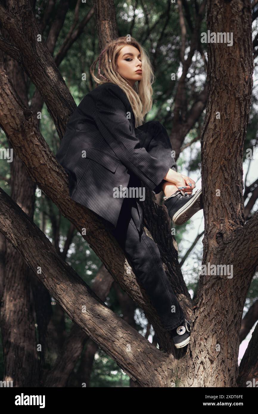 Eine junge Frau, ganz schwarz gekleidet, sitzt tagsüber auf einem Baum. Sie blickt von der Kamera weg, mit ihrem langen blonden Haar, das ihr Gesicht umrahmt. Stockfoto